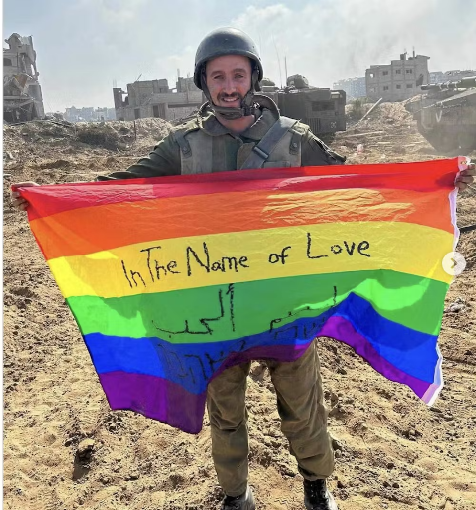 An Israeli Occupation Force soldier holding a rainbow pride flag. On the flag, "In the name of love" is written by hand in English, Arabic and Hebrew. The soldier stands in fron of the ruins of Gaza, in the background we see the rubble of carpet-bombed buildings.